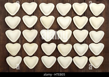 Rohen Teig in der Form eines Herzens (Knödel, Ravioli, Pelmeni), in Reihen auf einem dunklen hölzernen Hintergrund angelegt. Stockfoto