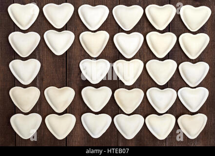 Rohen Teig in der Form eines Herzens (Knödel, Ravioli, Pelmeni), in Reihen auf einem dunklen hölzernen Hintergrund angelegt. Stockfoto