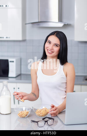 Attraktives Mädchen mit langen Haaren in die Kamera Lächeln und Löffel in der hand halten. Frau im weißen Oberteil Essen ihr Frühstück aus Eiche Flocken und Milch. In der Nähe von auf ta Stockfoto