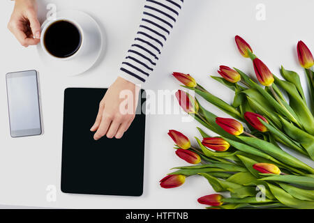 Frau Hände mit Tablet-Computer, Tasse Kaffee, Handy und schönen Blumen Haufen auf der weißen Bürotisch. Ansicht von oben Stockfoto