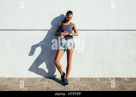 Voller Länge Aufnahme Fitness Frau stehend gegen eine Wand Fitness Fortschritte bei ihrer Smartwatch überprüfen. Fit weibliche Läufer Entspannung nach dem Training zu übertreffen Stockfoto