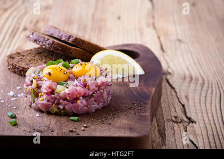 Steak Tartar in Ring-Form geformt, gekrönt mit zwei Wachteln Eigelb und mit Roggenbrot auf Holzbrett serviert Stockfoto