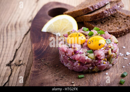 Steak Tartar in Ring-Form geformt, gekrönt mit zwei Wachteln Eigelb und mit Roggenbrot auf Holzbrett serviert Stockfoto