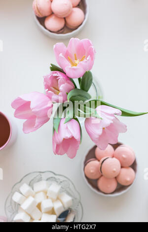 Frühling Hintergrund. Rosa Ma? Arons, Würfelzucker, eine Tasse Tee und einem Bouquet von rosa Tulpen auf einem weißen Tisch, Ansicht von oben. Stillleben mit frischen Bouquet von t Stockfoto