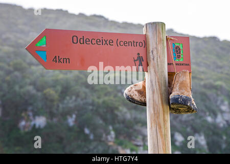 Wegweiser für Odeceixe, entlang der Rota Vicentina Weitwanderweg in der Algarve in Portugal, mit Wanderschuhen zum Trocknen gehängt. Stockfoto