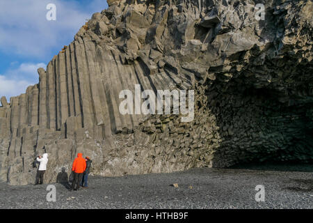 Verkleidet in warmen Jacken mit Kapuzen an einem windigen Tag bewundern die vulkanische sechseckige Basalt Felswand an Reynisfjara, Island, im Winter Stockfoto