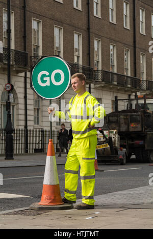 Ein gut sichtbarer Straßenarbeiter mit einem grünen „GO“-Schild, der den Verkehr in London leitet Stockfoto