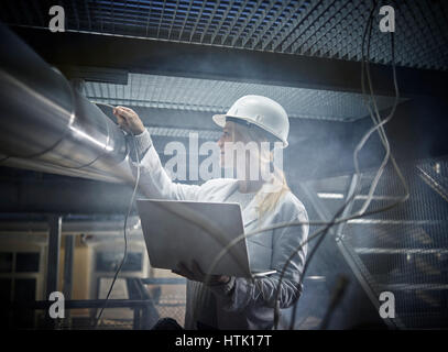 Junge Frau mit Helm, weißen Laborkittel und Laptop stand vor einer Industrieanlage, Österreich Stockfoto