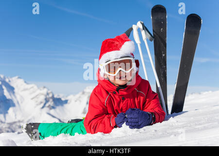 Niedliche kleine Skifahrer in Nikolausmütze Verlegung auf Schnee mit Ski und Stöcke in der Bank zu zwingen Stockfoto