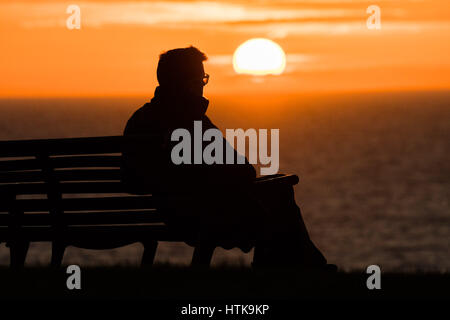 Aberystwyth Wales UK, Sonntag 12. März 2017 UK Wetter: ein Mann sitzt allein und starrt auf die untergehende Sonne spektakulär über den Wassern der Cardigan Bay aus Aberystwyth an der Westküste von Wales. Foto Credit: Keith Morris/Alamy Live-Nachrichten Stockfoto