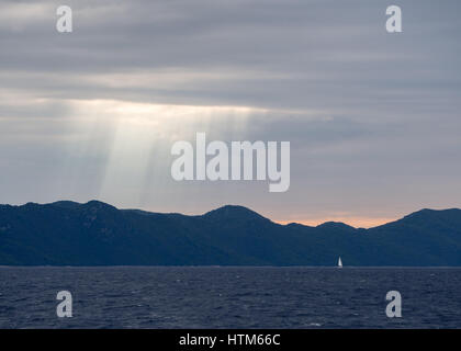 Sonnenstrahlen durch stürmische dunkle Wolken über eine Silhouette von Hügeln und Bergen Berg bei Sonnenuntergang mit Yacht am Meer Ozean, Kroatien Stockfoto