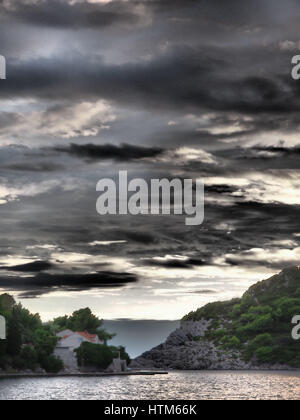 Eine böse stürmischer Sturm dunklen Himmel mit Wolken über eine felsige Insel Felsvorsprung mit Bäumen und roten Dach Haus, Sipan Kroatien Stockfoto