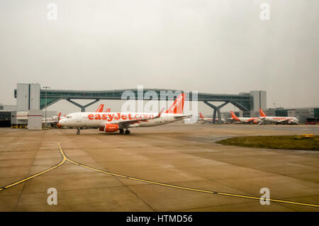 London Gatwick. England UK. EasyJet Flugzeugen am Flughafen London Gatwick Start- und Landebahn mit einem Fuß Brücke, die die Start- und Landebahn im Hintergrund überquert. Es gibt Flughafen Gebäude auf beiden Seiten der Landebahn, sowie mehr Flugzeuge am Terminal Tore geparkt. Stockfoto