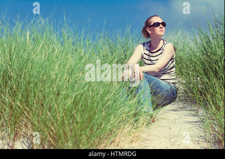 Junge Frau sitzt in Sanddünen unter hohe Gräser, entspannen, genießen den Blick auf sonnigen Tag, Luskentyre, Isle of Harris, Schottland Stockfoto