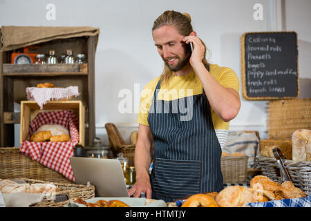Lächelnd männlichen Mitarbeiter telefonieren mit Handy während der Arbeit am Laptop am Schalter im Bäckerladen Stockfoto