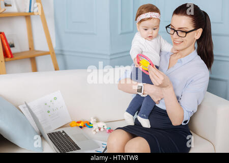 Bezaubernde Momente. Liebevolle sanfte schöne Mutter präsentieren ihre Tochter mit einem niedlichen rosa Spielzeug bei einer Rast für ein paar Minuten während ihrer Arbeitszeit Stockfoto