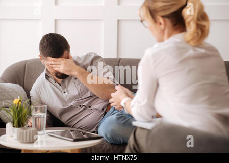 Während der psychologischen Sitzung. Depressiv düstere unglücklichen Mann hält seine Stirn und zum Nachdenken über seine persönlichen Probleme beim haben einer sozialmedizinischen Stockfoto