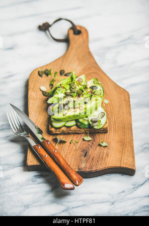 Gesundes Grün vegetarische Frühstück Konzept. Sandwich mit frischen Avocado, Gurke, Kohl, Kress sprießen, Kürbiskerne auf rustikalem Holzbrett über Marmor ba Stockfoto