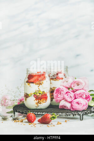 Gesund Frühling Frühstück. Griechischer Joghurt, Müsli, frische Erdbeer Frühstück in Gläser, rosa Raninkulus Blumen, Marmor Hintergrund, Tiefenschärfe, kopieren Stockfoto