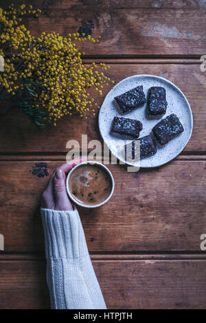 Zarte Frauenhand hält eine Tasse Kaffee auf einem rustikalen Holztisch, Platte mit Chocolat Coconut bars Stockfoto