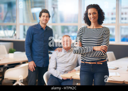 Porträt einer lächelnden Geschäftsfrau stehen selbstbewusst mit ihren Armen gekreuzt in einem modernen Büro mit zwei männlichen Kollegen im Hintergrund Stockfoto