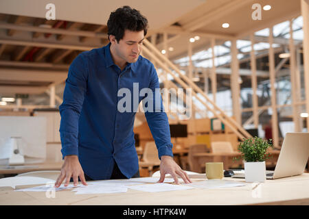 Lässig gekleidet fokussierten jungen Geschäftsmann, beugte sich über einen Schreibtisch in einem modernen Büro mit einem Laptop und Sortierung durch Papierkram Stockfoto