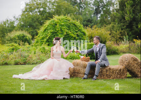 Klirrende Gläser. Braut und Bräutigam machen Toast sitzt auf einem Ballen Heu. Rasen, Bäume und Büsche im Hintergrund. Stockfoto