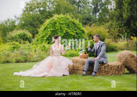 Braut und Bräutigam machen Toast sitzt auf einem Ballen Heu. Rasen, Bäume und Büsche im Hintergrund. Stockfoto