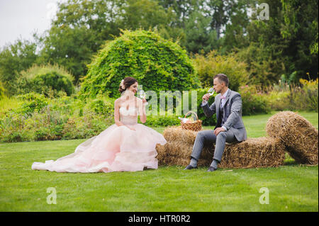 Klirrende Gläser. Braut und Bräutigam trinken Wein sitzen auf einem Ballen Heu. Rasen, Bäume und Büsche im Hintergrund. Stockfoto