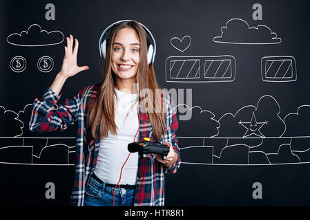 Verbringen Sie Zeit mit Vergnügen. Fröhlich lächelnde junge Frau tragen Kopfhörer und halten Spielkonsole beim Spielen von Videospielen Stockfoto