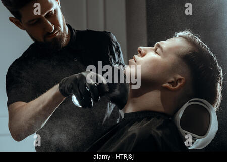 BARBERSHOP-THEMA. BÄRTIGE BARBER IN SCHWARZE GUMMIHANDSCHUHE GILT TALK MIT DEM PINSEL AUF CLIENT HAUT Stockfoto