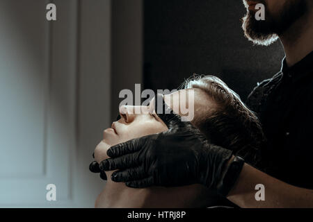 BARBERSHOP-THEMA. BÄRTIGE BARBER IN SCHWARZE GUMMIHANDSCHUHE GILT NACH RASIERGEL AUF CLIENT HAUT Stockfoto