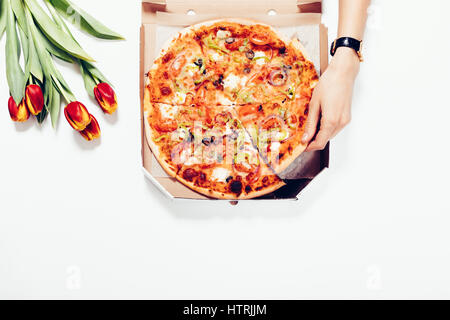 Zarte Frauenhand nimmt ein Stück Pizza aus dem Karton auf weißem Hintergrund, Ansicht von oben Stockfoto