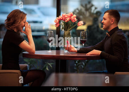 Junger Mann macht Frau Heiratsantrag und bietet ihr Schmuck bei Verlobungsring. Sie sitzen am Tisch neben Gläser Wein und Vase w Stockfoto