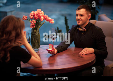 Junger Mann macht Frau Heiratsantrag und bietet ihr Schmuck bei Verlobungsring. Sie sitzen am Tisch neben Gläser Wein und Vase w Stockfoto