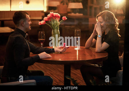 Junger Mann macht Frau Heiratsantrag und bietet ihr Schmuck bei Verlobungsring. Sie sitzen am Tisch neben Gläser Wein und Vase w Stockfoto