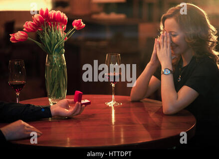 Junger Mann macht Frau Heiratsantrag und bietet ihr Schmuck bei Verlobungsring. Sie sitzen am Tisch neben Gläser Wein und Vase w Stockfoto