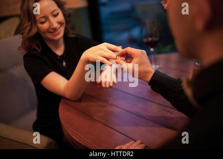 Junger Mann macht Frau Heiratsantrag und setzt ein Engagement ring an ihrem Finger. Sie sitzen am Tisch neben Gläser Wein. Stockfoto