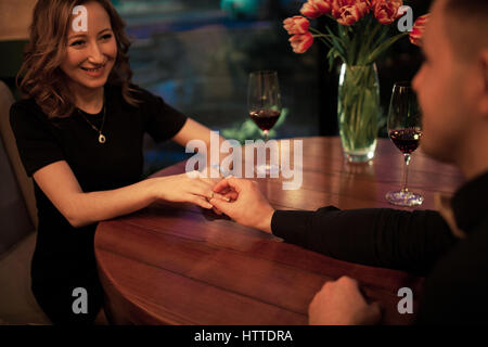 Junger Mann macht Frau Heiratsantrag und setzt ein Engagement ring an ihrem Finger. Sie sitzen am Tisch neben Gläser Wein und Vase mit fl Stockfoto