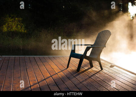 Entleeren Sie Adirondack Stuhl auf hölzerne Dock bei Sonnenaufgang mit Nebel rollt das Wasser. Stockfoto