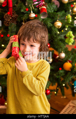 Kleine Junge schüttelte ein Geschenk nah an seinem Ohr versuchen zu erraten, was es ist. Er steht vor dem Weihnachtsbaum. Stockfoto