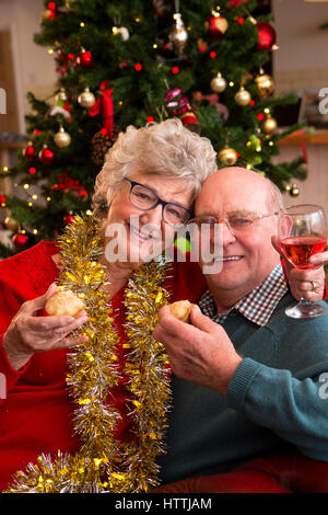 Älteres paar posiert für die Kamera zu Weihnachten. Sie halten Mince Pies vor dem Weihnachtsbaum. Stockfoto