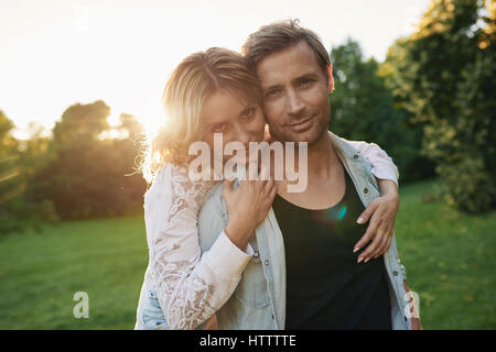 Porträt einer lächelnden jungen Frau umarmt ihr Mann von hinten stehen zusammen in einem Park am späten Nachmittag Stockfoto