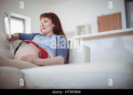 Schwangere Frau mit Kopfhörern auf den Bauch entspannen Sie sich auf sofa Stockfoto