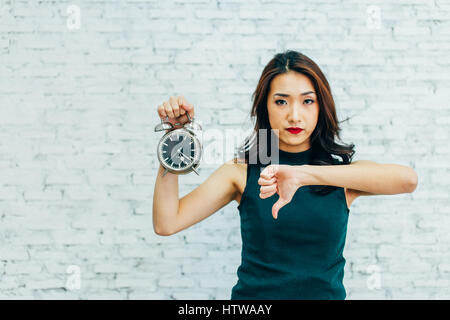 Asian Business Frau, Daumen nach unten und halten Wecker - was darauf hinweist, dass nicht mit der Zeit Management zufrieden Stockfoto