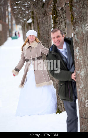 Braut steht hinter Bräutigam vom Baum im Winter Park herausschauen, Spaziergänge Brautpaar Stockfoto