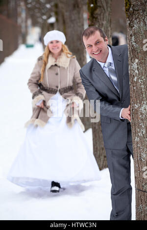 Listige Braut Greeping bis der Bräutigam spähen aus Baum im Winter park Stockfoto