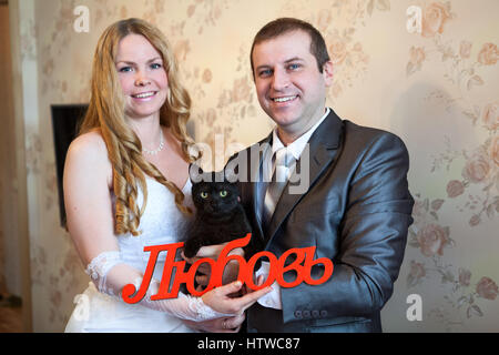 Freudige kaukasischen Hochzeitspaar halten rote Wort Liebe auf Russisch in Händen und schwarze Katze Stockfoto