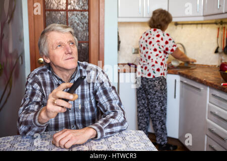 Kaukasische senior woman schaltet Sender mit TV-Fernbedienung beim Sitzen am Tisch in der Küche, Frau waschen utensil Stockfoto