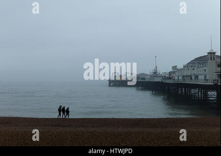 Drei junge Leute, die zu Fuß am Strand im frühen Morgennebel durch Brighton Pier Stockfoto